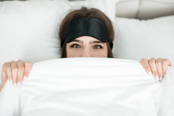 jeune femme attirante avec le masque de sommeil sur sa tête se trouvant dans le lit sous la couverture le matin - waking up women bedding underwear photos et images de collection