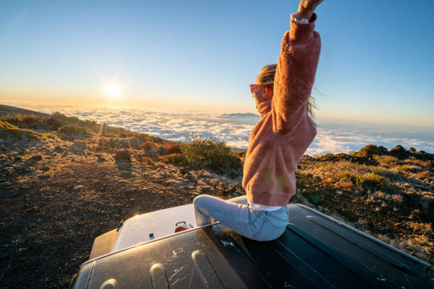 istock Young woman sitting on car arms open at sunset above clouds 1193233064