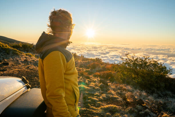 Young man standing near vehicle on a volcano and looking at sun setting over the cloudscape