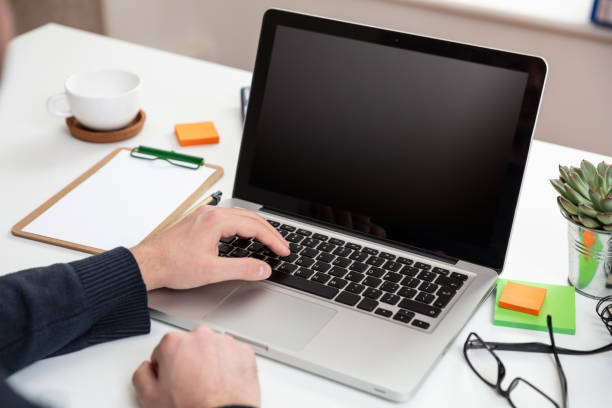 Computer screen mockup. Man working with a black blank screen laptop on a wood desk, office background, copy space