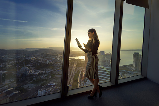 Young woman standing in front of large window in a business office during sunset.