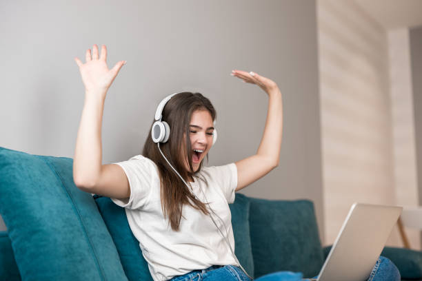 urocza młoda kobieta w słuchawkach słuchanie muzyki z laptopa i granie w gry wideo na kanapie w salonie wygrał quest - rythm zdjęcia i obrazy z banku zdjęć