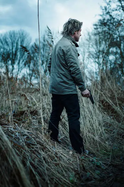 Photo of Man with pistol between reed on cloudy day.