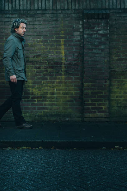 istock Man in green jacket walking on pavement along brick wall. 1193259125