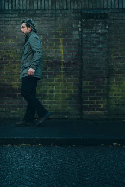 Man in green jacket walking on pavement along brick wall.