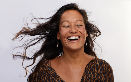 Mature woman with long tousled brown hair laughing with her eyes closed while standing against a gray background