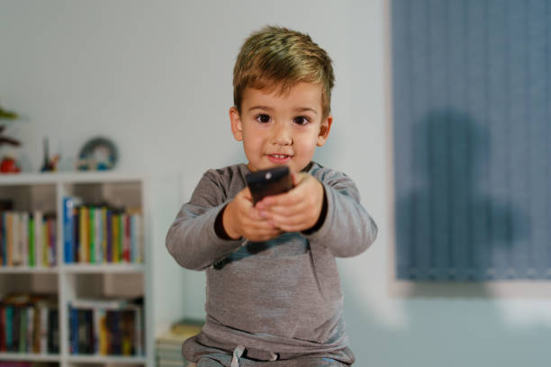 portrait of young small boy sitting on the bed at home holding remote controller at home three years old caucasian cute watching tv television - boyhood imagens e fotografias de stock