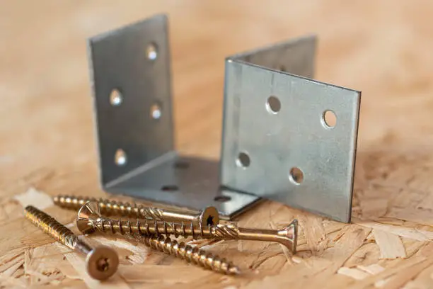 Two galvanized angle brackets and self drilling screws lying on chip board. Blurred background.