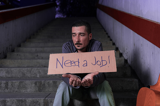 View of Unemployed Person with Lot of Life Problems who is Sitting on the Stairways in the Subway with Donation Box and Paper Cardboard. Sad Homeless Young Man or Beggar is Asking for Money Donation. Poverty and Social Issue Concept.