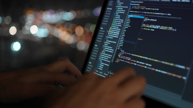 The programmer writes the code for the development of the website, against the background of a beautiful night window in which the city lights are visible in defocus.