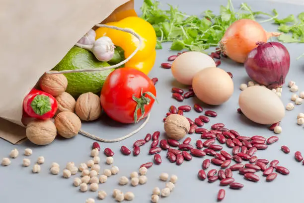 Photo of A set of products for healthy eating. Paper bag with walnuts, chickpeas, beans, lentils, yellow and red peppers, tomatoes, avocado on a gray background. Organic food. Top view