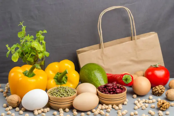 Photo of A set of products for healthy eating. Paper bag with walnuts, chickpeas, beans, lentils, yellow and red peppers, tomatoes, avocado on a gray background.