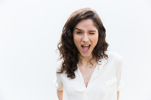 Excited cheerful student girl winking at camera. Wavy haired young woman in casual shirt standing isolated over white background. Communication concept