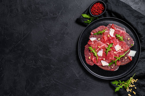 Marbled beef carpaccio with arugula and parmesan cheese. Black background. Top view. Space for text Marbled beef carpaccio with arugula and parmesan cheese. Black background. Top view. Space for text. carpaccio parmesan cheese beef raw stock pictures, royalty-free photos & images