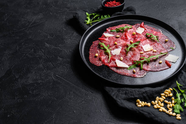 Marbled beef carpaccio with arugula and parmesan cheese. Black background. Top view. Space for text Marbled beef carpaccio with arugula and parmesan cheese. Black background. Top view. Space for text. carpaccio parmesan cheese beef raw stock pictures, royalty-free photos & images