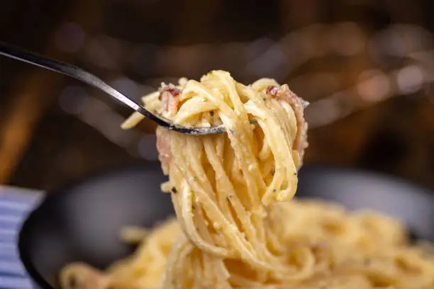 Spaghetti Carbonara with bacon is being lifted by a fork from a bowl before taking a bite