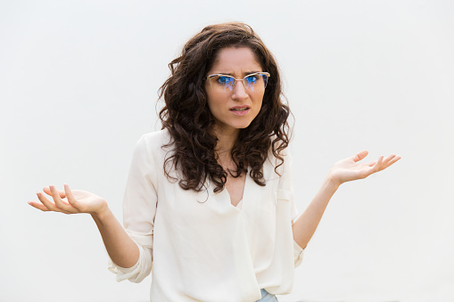 Puzzled annoyed woman in glasses shrugging and staring at camera. Wavy haired young woman in casual shirt standing isolated over white background. Asking question concept