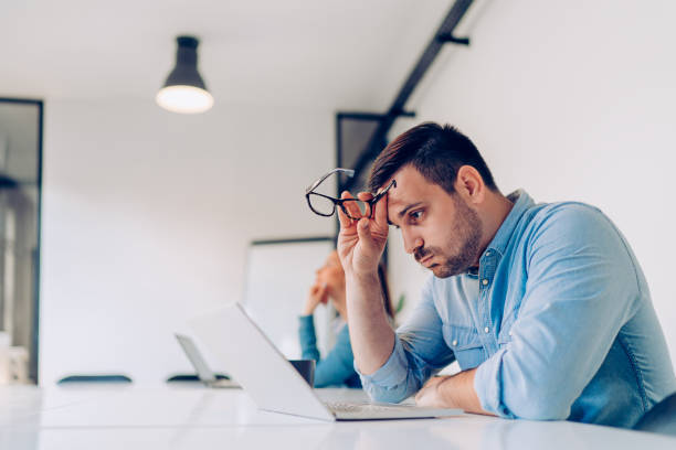Exhausted young businessman at work Exhausted young businessman using laptop at work and sitting by the desk while edgy man stock pictures, royalty-free photos & images