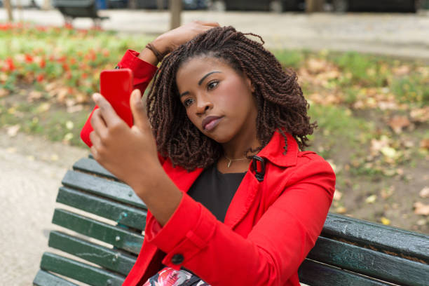 Pensive woman using smartphone in park. Beautiful thoughtful young African American woman sitting on bench and using mobile phone. Technology concept