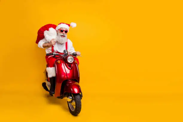 Full body photo of crazy funny santa claus in red hat drive hurry fast motor cycle hold sack, christmas x-mas preparation ride north-pole wear shirt suspenders isolated yellow color background