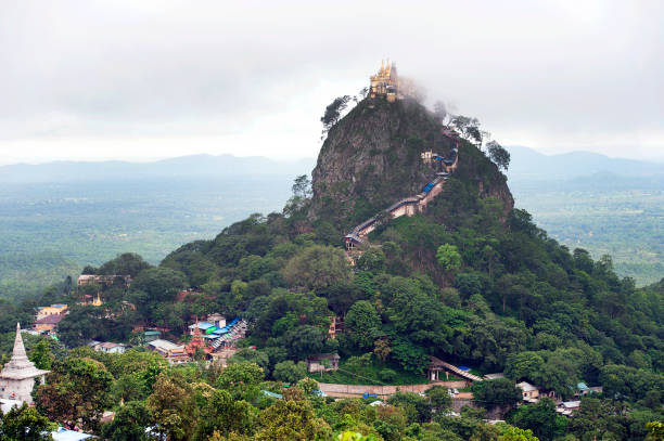 Mount Popa Buddhist temple stupa and buildings with meandering stairway of covered stone steps seen obscured in mist and cloud at the top peak of a rocky extinct volcano in central Myanmar in the region of Mandalay, 50 km southeast of Bagan in the Pegu Range. It can be seen from the Ayeyarwady River as far away as 60 km in clear weather