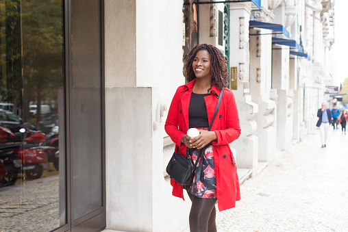 Smiling woman with coffee to go walking on street. Attractive young woman in red trench coat holding paper cup and walking near building. Fashion concept