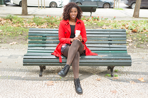 Woman with coffee to go smiling at camera. Full length view of cheerful young African American woman sitting on bench with paper cup. Hot drink concept