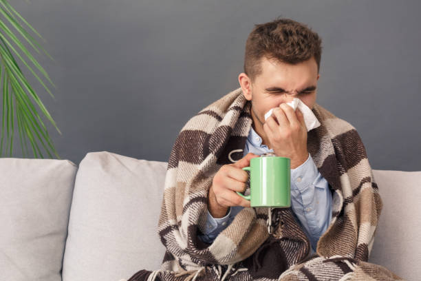 istock Feeling Sick. Young man sitting on sofa under blanket studio isolated on grey with cup of tea blowing his nose 1193321856