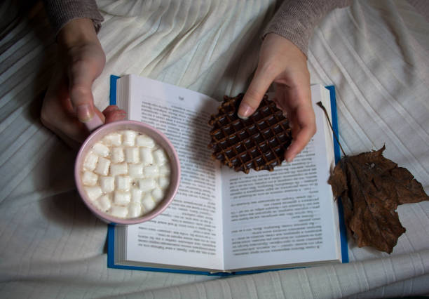 Girl drinks cocoa with marshmallow from pink cup with chocolate wafel and reads book