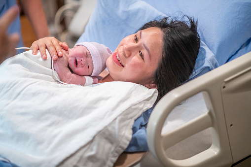 A young Asian mother lays in her hospital bed in the delivery room after giving birth to her son.  She has him laying on her chest as the share an intimate moment together and some skin-to-skin time.  She is wearing a blue hospital gown and has a white blanket draped over the newborn.
