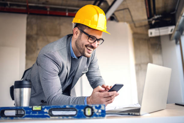 lächelnd attraktive kaukasische architekt mit helm auf dem kopf halten smartphone und blick auf laptop, während im gebäude im bauprozess stehen. - building contractor computer internet construction worker stock-fotos und bilder