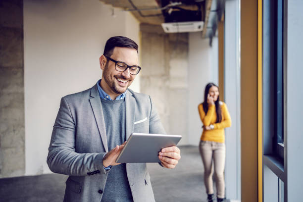 handsome immobilienmakler im anzug stehen im gebäude im bauprozess und mit tablet für die verabredung mit kunden. im hintergrund stand sein kollege und telefonierte. - business ceo building activity business person stock-fotos und bilder