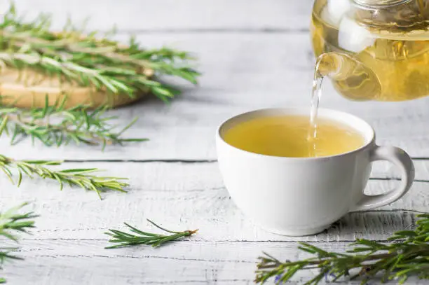 Photo of White cup of healthy rosemary tea pouring from teapot with fresh rosemary bunch on white wooden rustic background, winter herbal hot drink concept, salvia rosmarinus