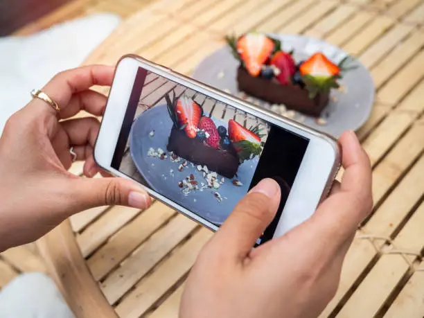 Photo of Woman's hand taking a photo of delicious chocolate tart on ceramic dish by smartphone in cafe.