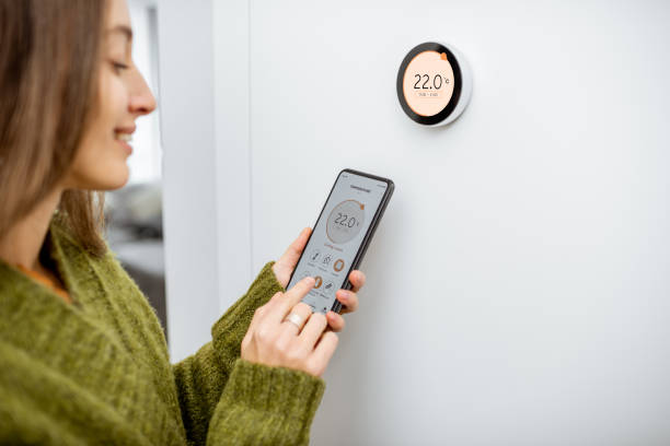 Woman regulating heating temperature with phone and thermostat at home Woman dressed in green sweater regulating heating temperature with a modern wireless thermostat and smart phone at home. Synchronization of thermostat with mobile devices concept smart thermostat stock pictures, royalty-free photos & images