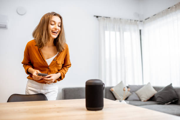 Woman controlling home devices with a voice commands Young cheerful woman controlling home devices with a voice commands, talking to a smart column at home. Concept of smart home and voice command control virtual assistant stock pictures, royalty-free photos & images