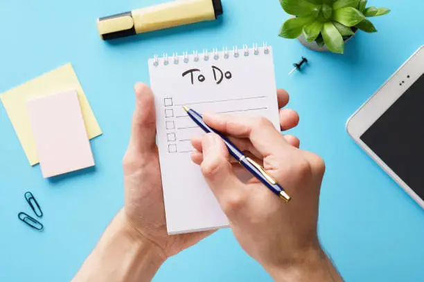 Male hands making a to-do list in a notebook over a blue office desk.
