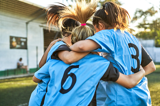 Female soccer players are huddling on sports field. Girls are celebrating success after winning match. They are in blue sportswear.