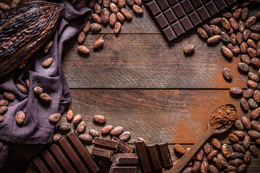 Top view of a frame made with some chocolate bars, cocoa seeds, cocoa powder and a cocoa pod on a dark brown wooden backdrop. There is a useful copy space for a text or a logo at the center of the image. Low key DSLR photo taken with Canon EOS 6D Mark II and Canon EF 24-105 mm f/4L