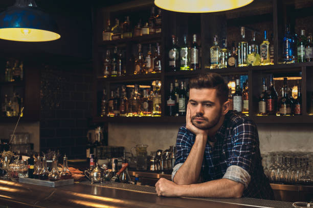 Young bartender leaning on bar counter thoughtful Young barman standing leaning on bar counter looking down thoughtful pub bar counter bar men stock pictures, royalty-free photos & images