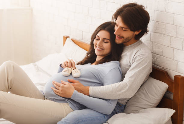 happy expecting couple lying on bed with little shoes for baby - abdomen women loving human hand imagens e fotografias de stock