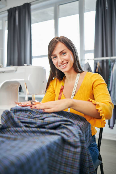 Smiling charming caucasian fashion designer sitting in her studio and sewing beautiful evening dress. stock photo