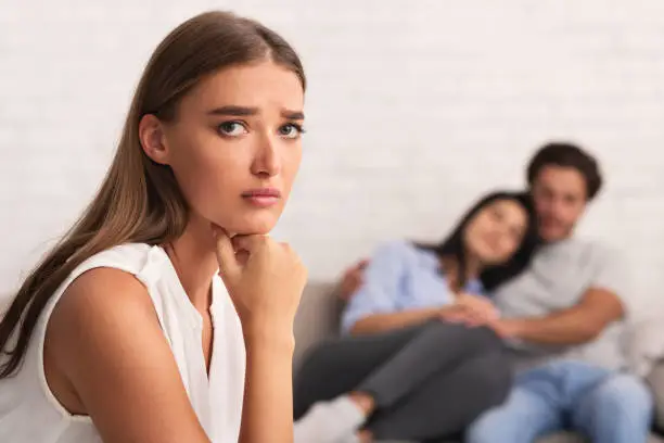 Jealousy. Sad Girl Looking At Camera While Her Ex-Boyfriend Hugging New Girlfriend Sitting On Couch Indoor. Selective Focus