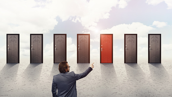 Businessman with glasses pointing at red door among other seven doors over sky background, opportunity concept