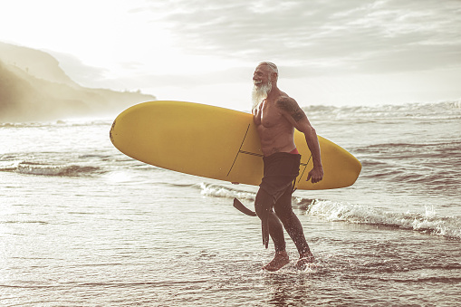 Senior fit guy walking with longboard after surfing at sunset - Mature tattooed man having fun doing extreme sport on tropical beach - Joyful elderly lifestyle and travel concept - Focus on male body