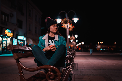 Casual dressed teenage girl sitting on the bench in the city street and drinking coffee on a winter night