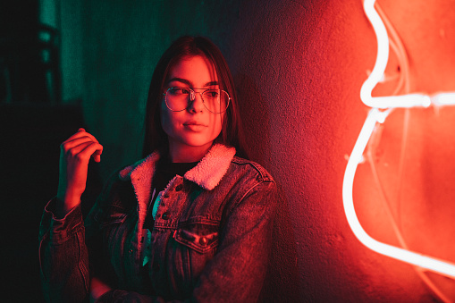 Beautiful casual dressed teenage girl standing next to a neon light on a night in the city