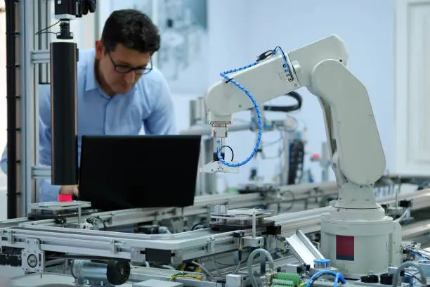 Photo of robotic arm which picks up product from automated car on production line and an engineer working on laptop at background