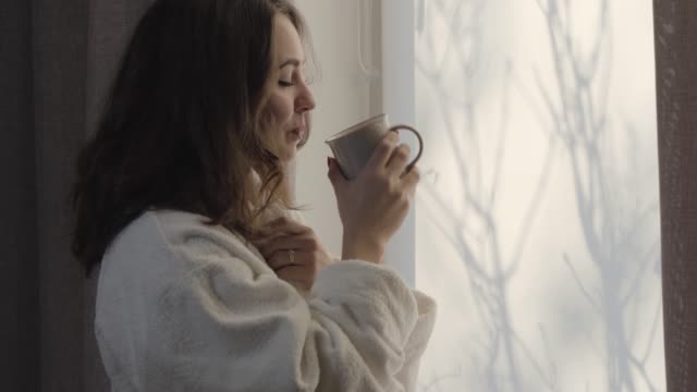 Portrait of cute Caucasian brunette girl drinking morning coffee and smiling. Gorgeous young brunette woman looking out the window. Lady in white bathrobe enjoying sunny day.