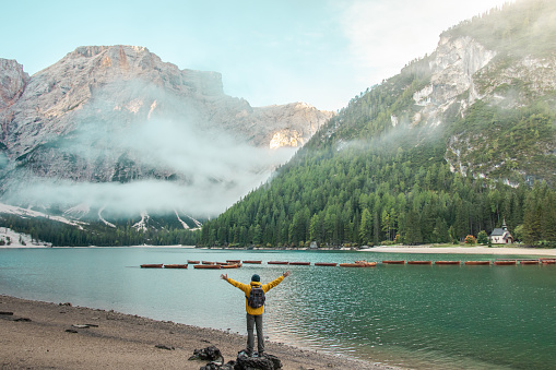 Young tourist hiker man at beautiful alpine lake Lago Di Braies (Pragser wildsee) in Trentino. Raising hands. Dolomites mountains, Italy.